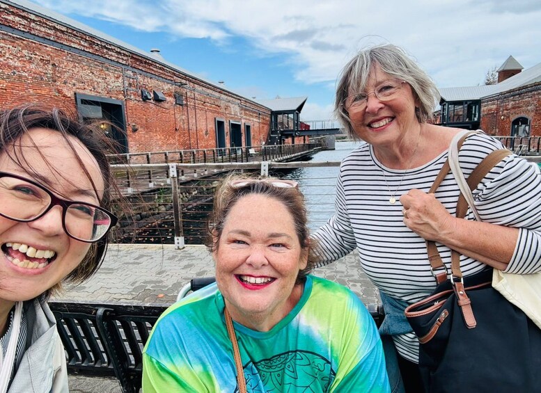 Three people sitting on a bench near a historic brick building with a waterway and a bridge in the background. 3