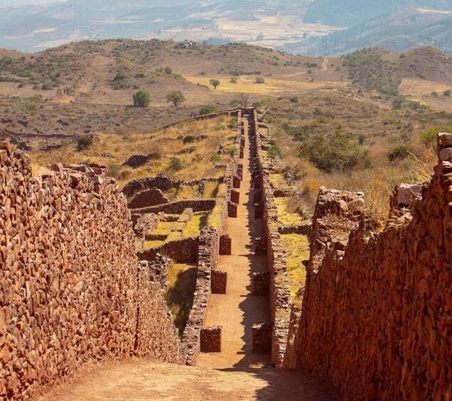 An ancient stone pathway leading through a dry, hilly landscape with ruins on either side. 1
