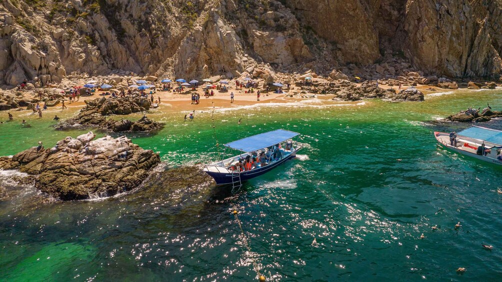 A boat on clear turquoise waters surrounded by rocky formations and a beach with people and umbrellas. 1