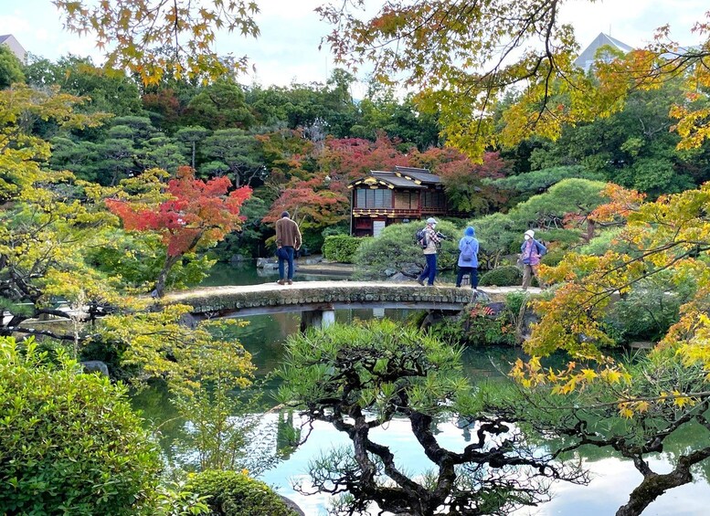 A serene garden with a pond, people walking, and traditional Japanese architecture. 1