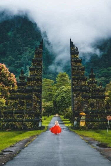 A person in a red dress walking down a path between two ancient temple structures surrounded by lush greenery and mist. 3