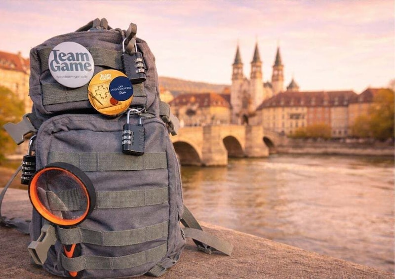 A backpack with a 'Team Game' patch and a ball, sitting by a river with a historic bridge in the background. 1