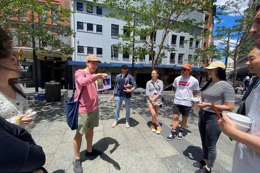 A group of people standing on a sidewalk, some holding food items, with buildings and trees in the background. 3