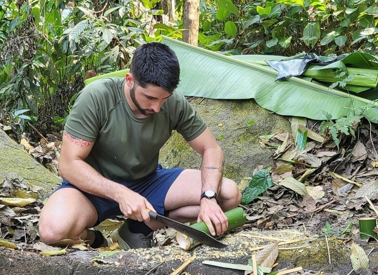 A man cutting a green plant with a knife in a forested area. 1