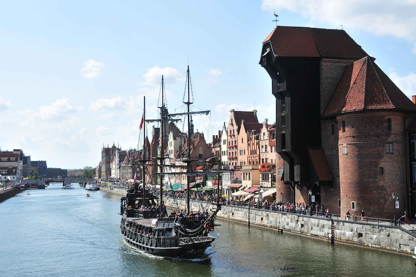A tall ship docked at a harbor with historic buildings and a bridge in the background. 2