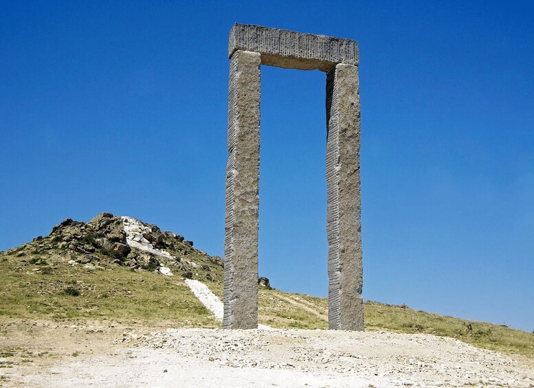 A tall, narrow stone structure with inscriptions, standing on a grassy hill under a clear blue sky. 5