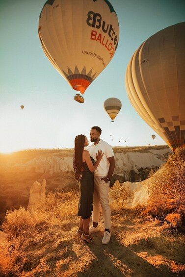 A couple standing on a hill with hot air balloons in the background. 1