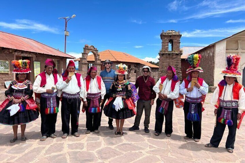 A group of people in traditional attire standing in a plaza with stone pavement, a church, and a bell tower in the background. 3