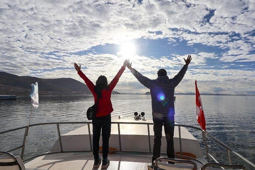 Two people on a boat with their arms raised, a flag, and a body of water with mountains in the background. 2