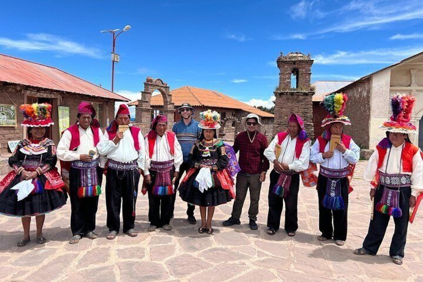 A group of people in traditional attire standing in a plaza with stone pavement, a church, and a bell tower in the background. 4