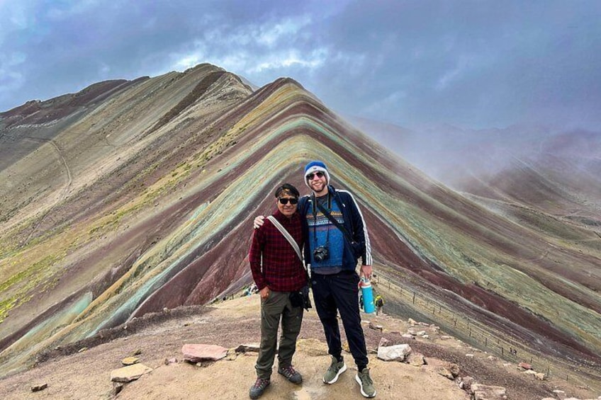 Two people standing on a mountain ridge with a colorful striated rock formation in the background. 3