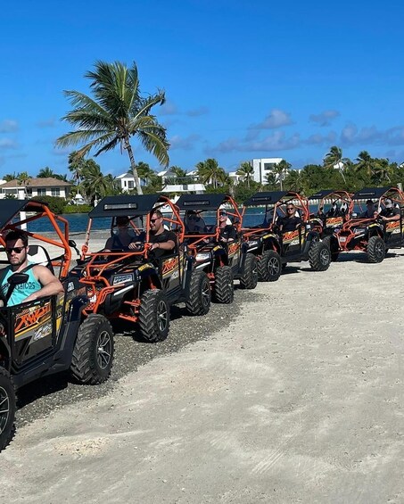 A row of orange off-road vehicles parked on a sandy beach with palm trees and houses in the background. 1