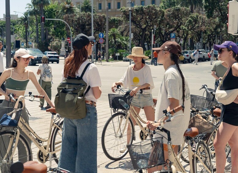 A group of people standing around bicycles on a sunny street with palm trees and cars in the background. 5