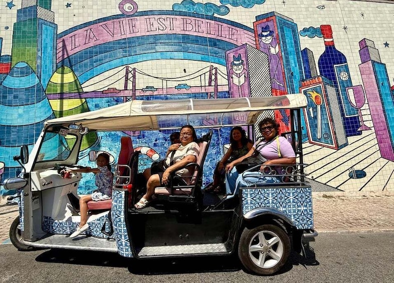A tuk-tuk with passengers in front of a colorful mural. 2