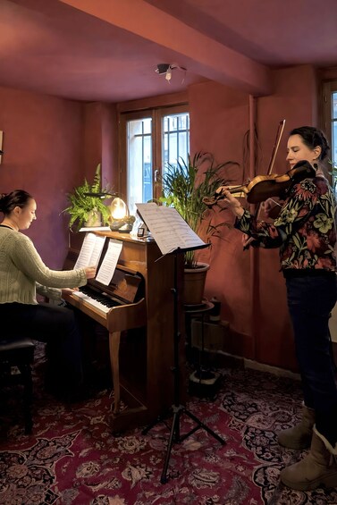 A person playing the piano and another playing the violin in a room with a piano, sheet music, and potted plants. 2