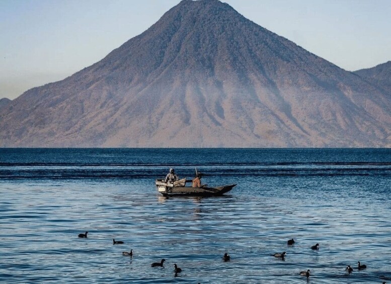 A boat with two people on a calm body of water with ducks swimming around. In the background, there is a large mountain. 3