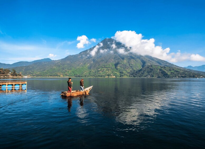 A calm lake with a wooden boat carrying two people, a mountain with clouds in the background, and a dock with barrels on the left. 2