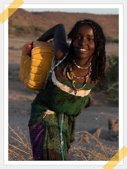 A person carrying a large yellow container on their back, wearing a green patterned dress and multiple necklaces, in a dry, open landscape. 1