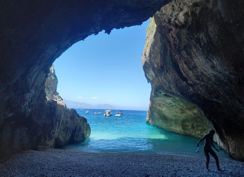 A rocky cave opening to a clear blue sea with boats and a person on the shore. 1