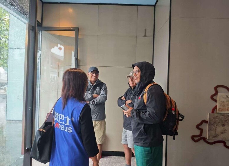 A group of people standing in a glass-walled entrance area, some wearing casual clothing and backpacks. 2