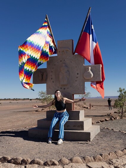 A person standing in front of a stone monument with two flags, one multicolored and one red and white, in a desert landscape. 3