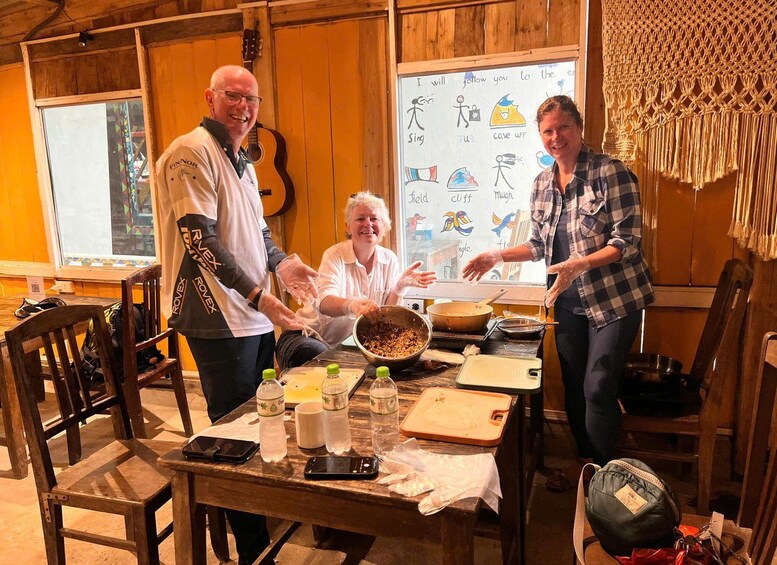 Three people in a rustic kitchen preparing food, with a guitar and a wall with drawings in the background. 2