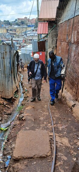 Two men walking down a narrow alley with corrugated metal walls, a drainage ditch, and a hose on the ground. 1
