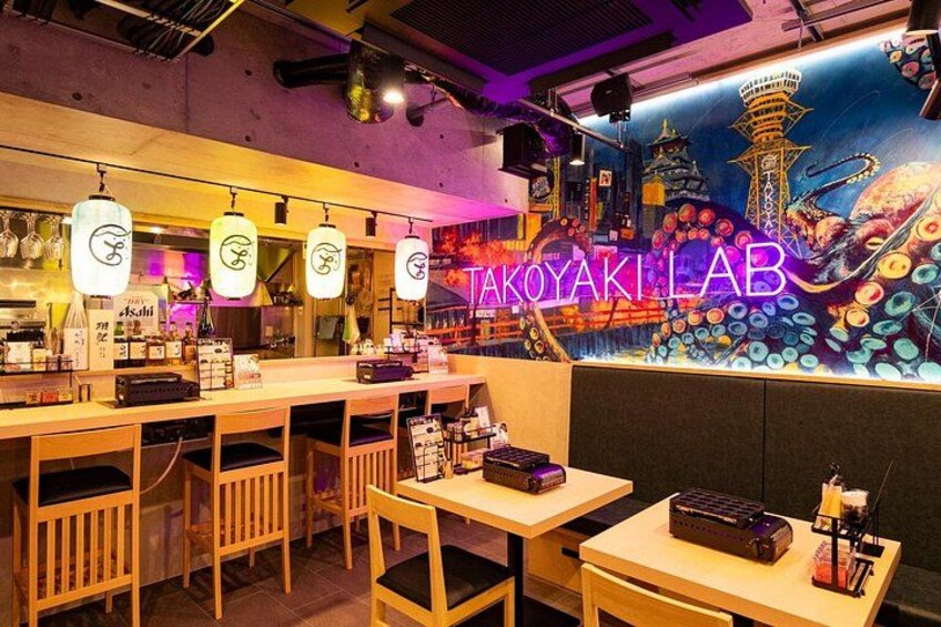 L’intérieur d’un restaurant, avec une enseigne au néon indiquant « TAKOYAKI LAB », des meubles en bois et une fresque représentant un paysage urbain. 4