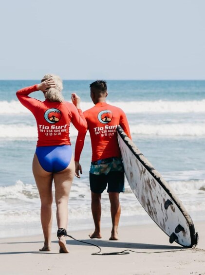 Two people in red wetsuits with 'Tio Surf' logos walking on a beach with a surfboard. 4