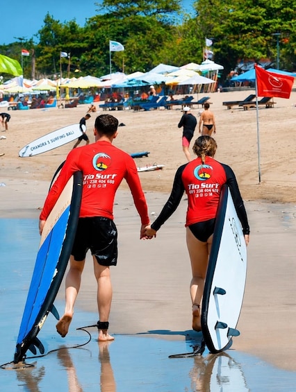 Two people in red wetsuits carrying surfboards on a beach. 1