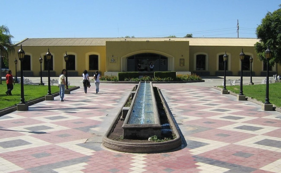 A courtyard with a central water feature, a yellow building with arched windows, and people walking. 3