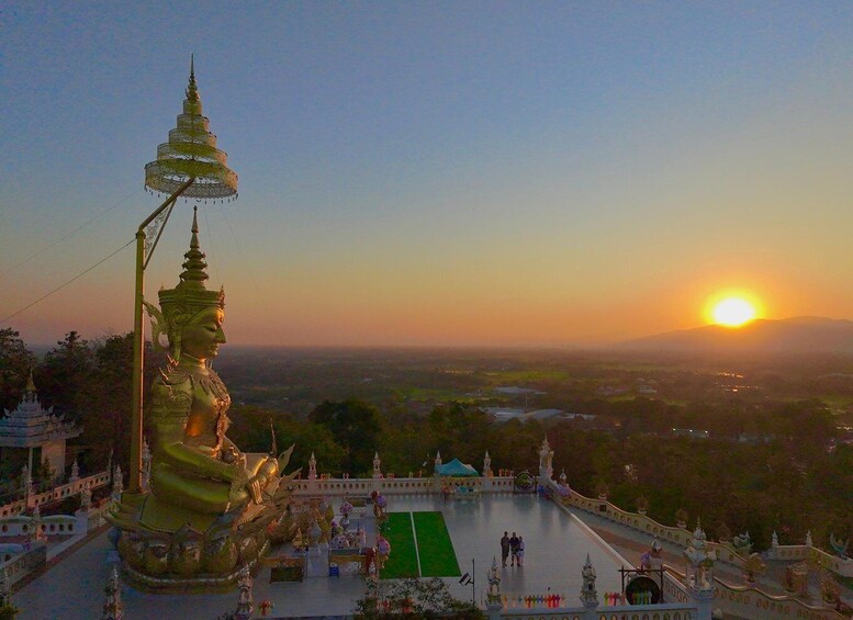 Een tempelcomplex met een groot gouden beeld, een pagode en een ondergaande zon op de achtergrond. 1