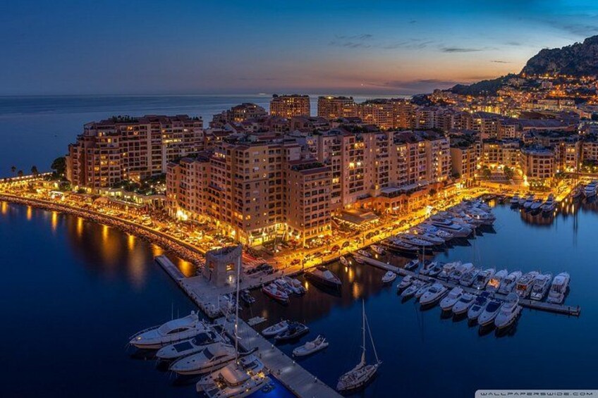 Eine Küstenstadt im Abendlicht mit beleuchteten Gebäuden, einer Marina mit Booten und ruhiger See. 2