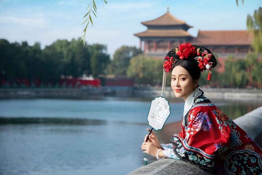 A person in traditional attire holding a fan, sitting by a body of water with a pagoda in the background. 2