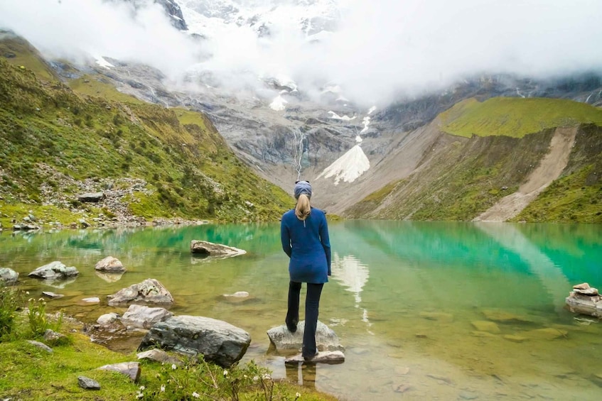 A person standing on rocks by a clear turquoise lake surrounded by mountains and snow. 3