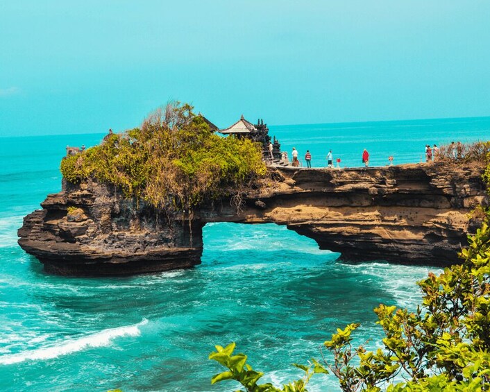 A rock formation with a natural arch over turquoise waters, a small structure on the rock, and people walking along the edge. 1