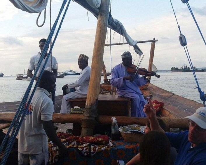 Eine Gruppe von Menschen auf einem Boot, einige spielen ein Saiteninstrument, im Hintergrund ruhiges Meer und weitere Boote. 3