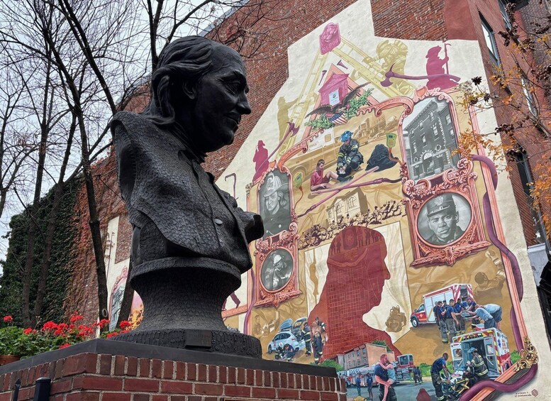 A statue of a man with a bust base, a mural with firefighters, and a building in the background. 1