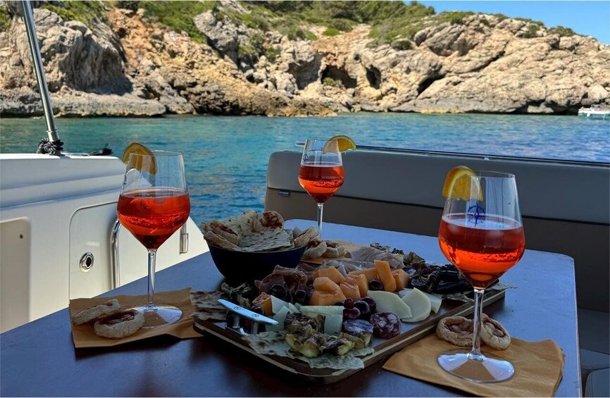 A table with a charcuterie board and two glasses of red wine on a boat with a rocky coastline in the background. 2