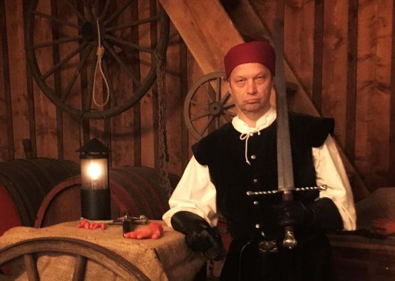 A person in historical attire holding a sword, with wooden barrels and a lantern in the background. 1