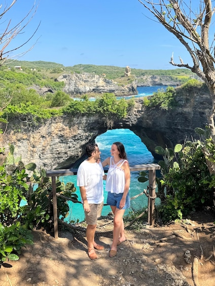 A couple standing by a cliff overlooking a turquoise sea with a natural rock arch formation. 2