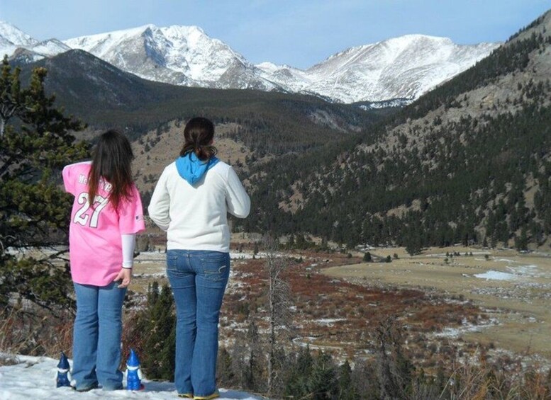 Dos personas están de pie en una montaña nevada, con vistas a un valle y montañas cubiertas de nieve al fondo. 3