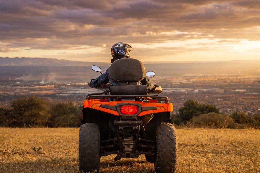 Una persona conduce un cuatrimoto naranja sobre una colina cubierta de hierba, con vistas a una ciudad al atardecer. 3