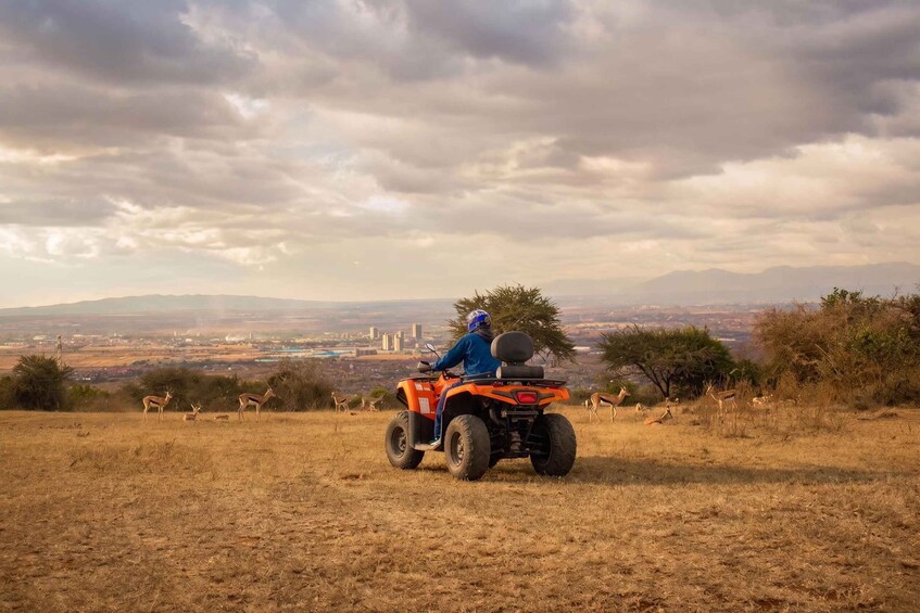 Una persona conduciendo un cuatrimoto naranja en una colina cubierta de hierba, con un paisaje urbano al fondo. 2