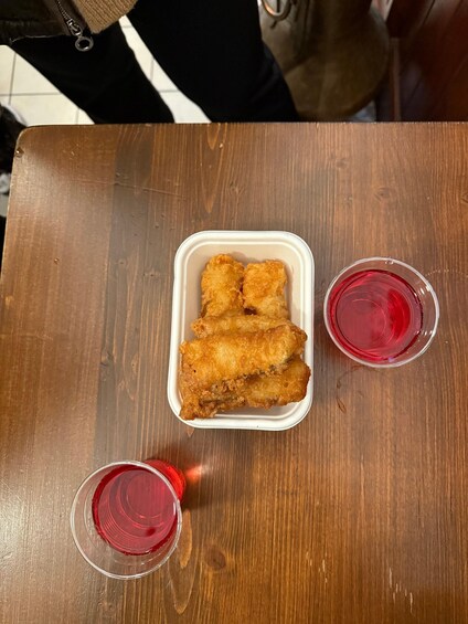 A wooden table with a white takeout container holding fried chicken, two glasses of red beverage, and a partial view of a person. 3