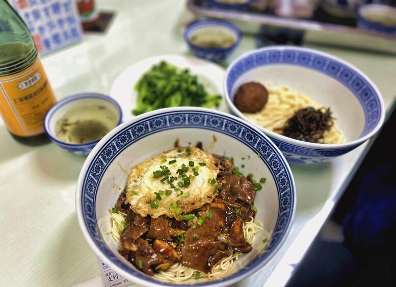 A bowl of noodles topped with a fried egg and green onions, with a bottle and other dishes in the background. 4