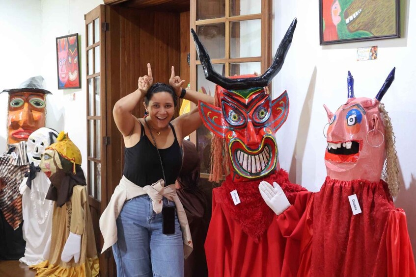 A person posing with traditional masks in a room with wooden doors and framed artwork on the walls. 4