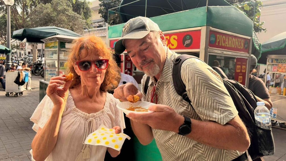 Two people eating food at an outdoor market with a food cart labeled 'PICARONES' in the background. 1