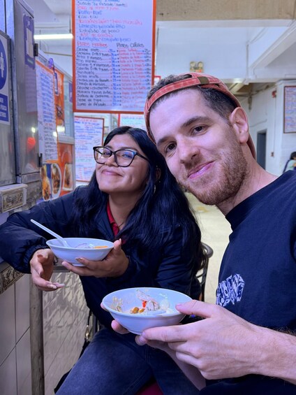 Two people sitting at a table with food, in a restaurant with menus on the wall. 3