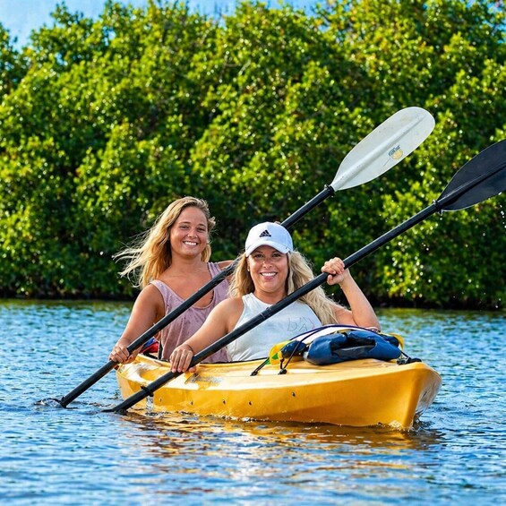 Two people kayaking on a yellow raft in calm water with dense green foliage in the background. 1